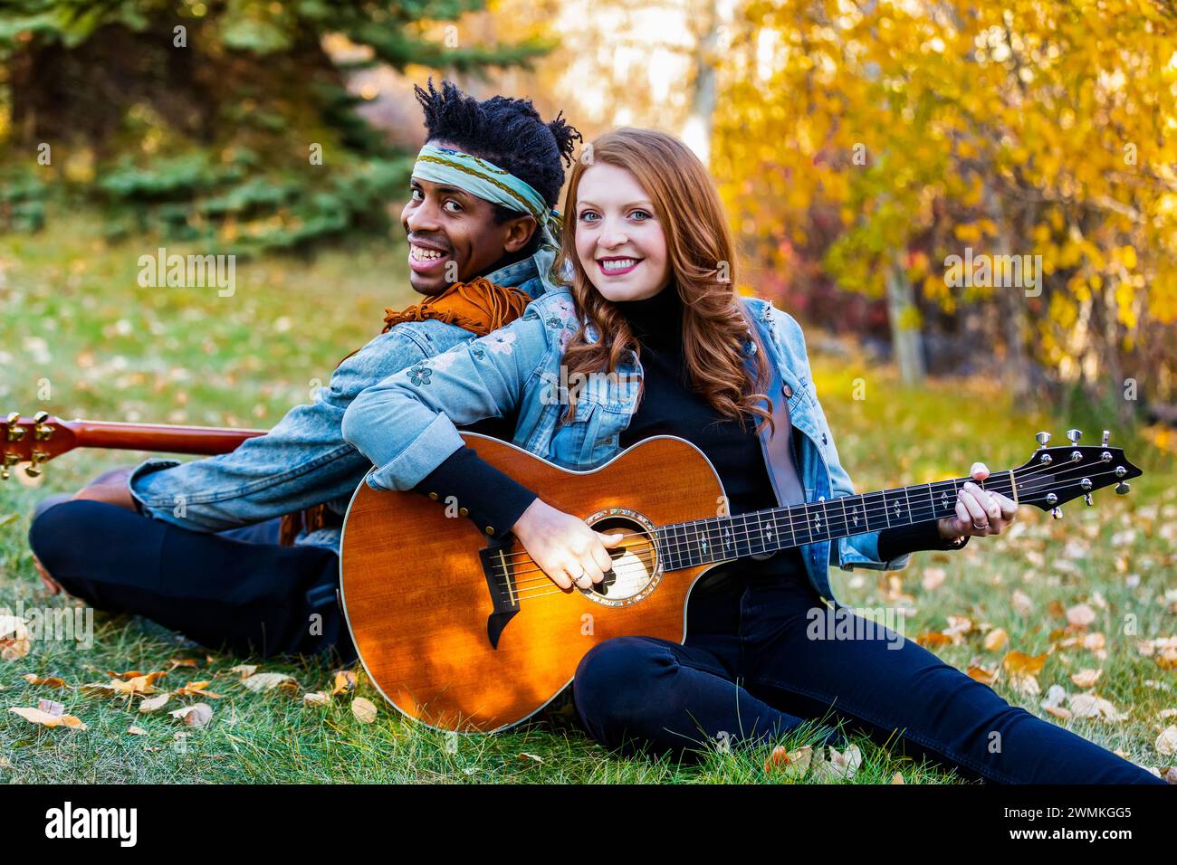 Portrait rapproché d'un couple marié de race mixte assis sur l'herbe dos à dos jouant de la guitare, souriant et posant pour la caméra pendant une chute... Banque D'Images