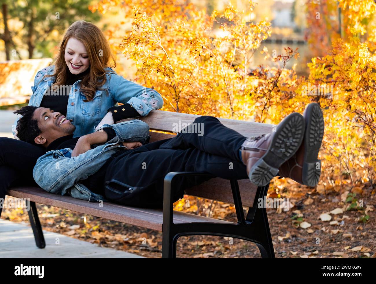 Un couple de courses mixtes souriant l'un à l'autre et se relaxant sur un banc de parc tout en passant du temps de qualité ensemble lors d'une sortie en famille d'automne dans un parc de la ville Banque D'Images