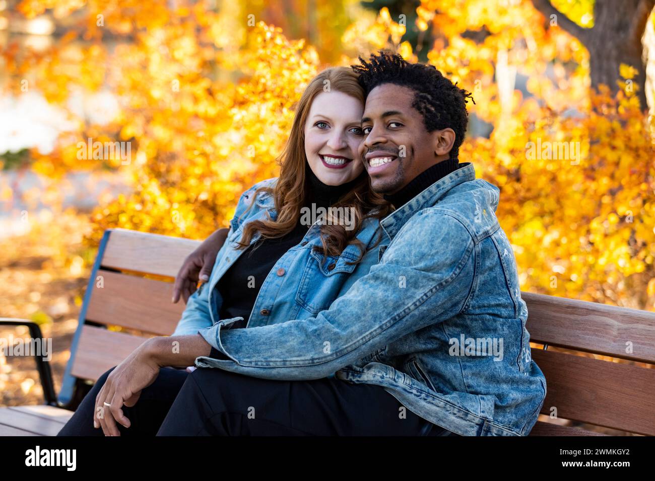 Portrait d'un couple de races mixtes embrassant et souriant devant la caméra, assis sur un banc de parc, passant du temps de qualité ensemble pendant une famille d'automne... Banque D'Images