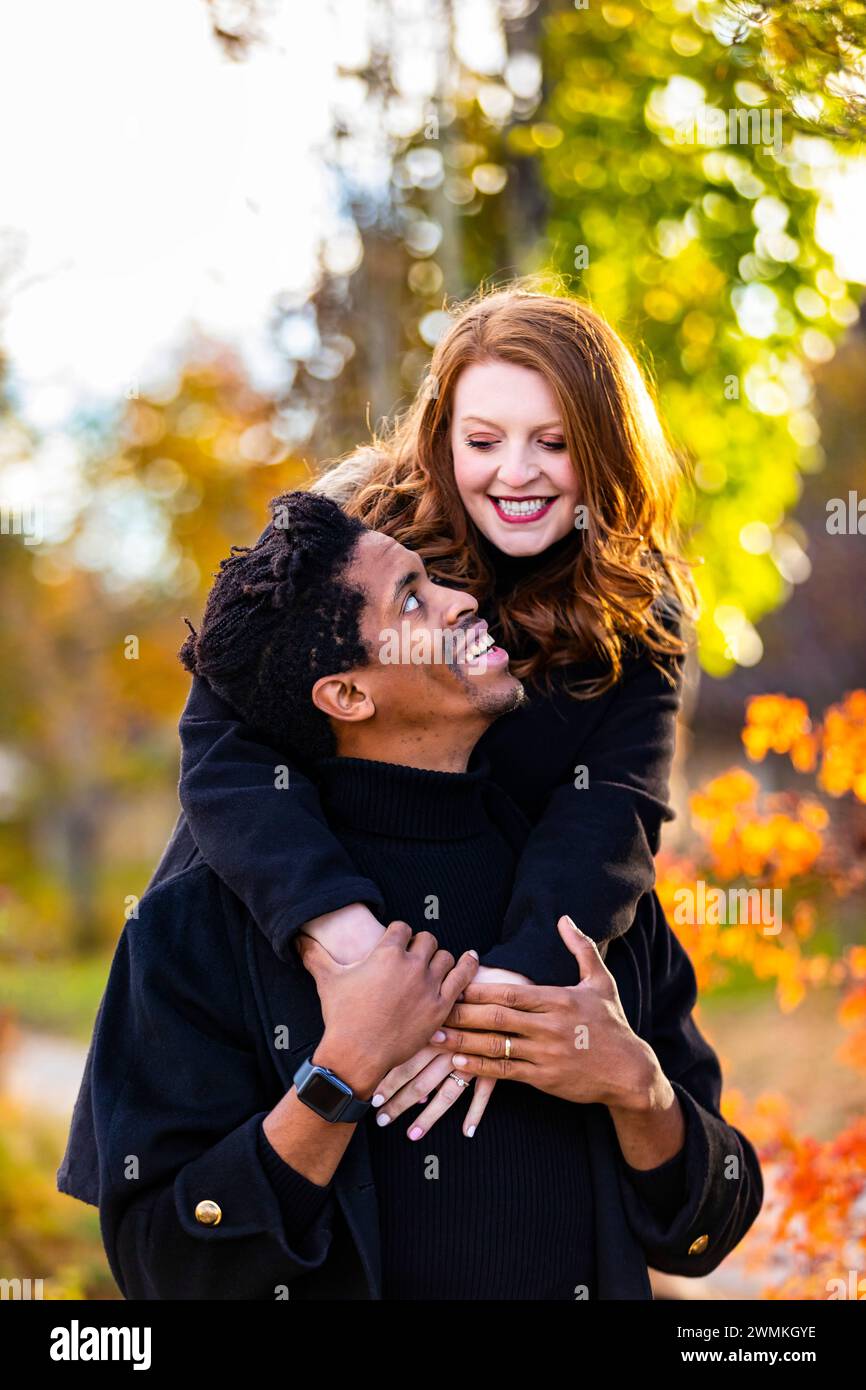 Portrait d'un couple de races mixtes, épouse serrant son mari par derrière, tout en passant du temps de qualité ensemble lors d'une sortie en famille d'automne dans un parc de la ville Banque D'Images