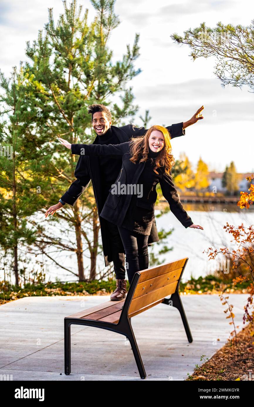 Portrait d'un couple de races mixtes marchant sur un banc de parc, en équilibre et souriant à la caméra, passant du temps de qualité ensemble lors d'une famille d'automne o... Banque D'Images