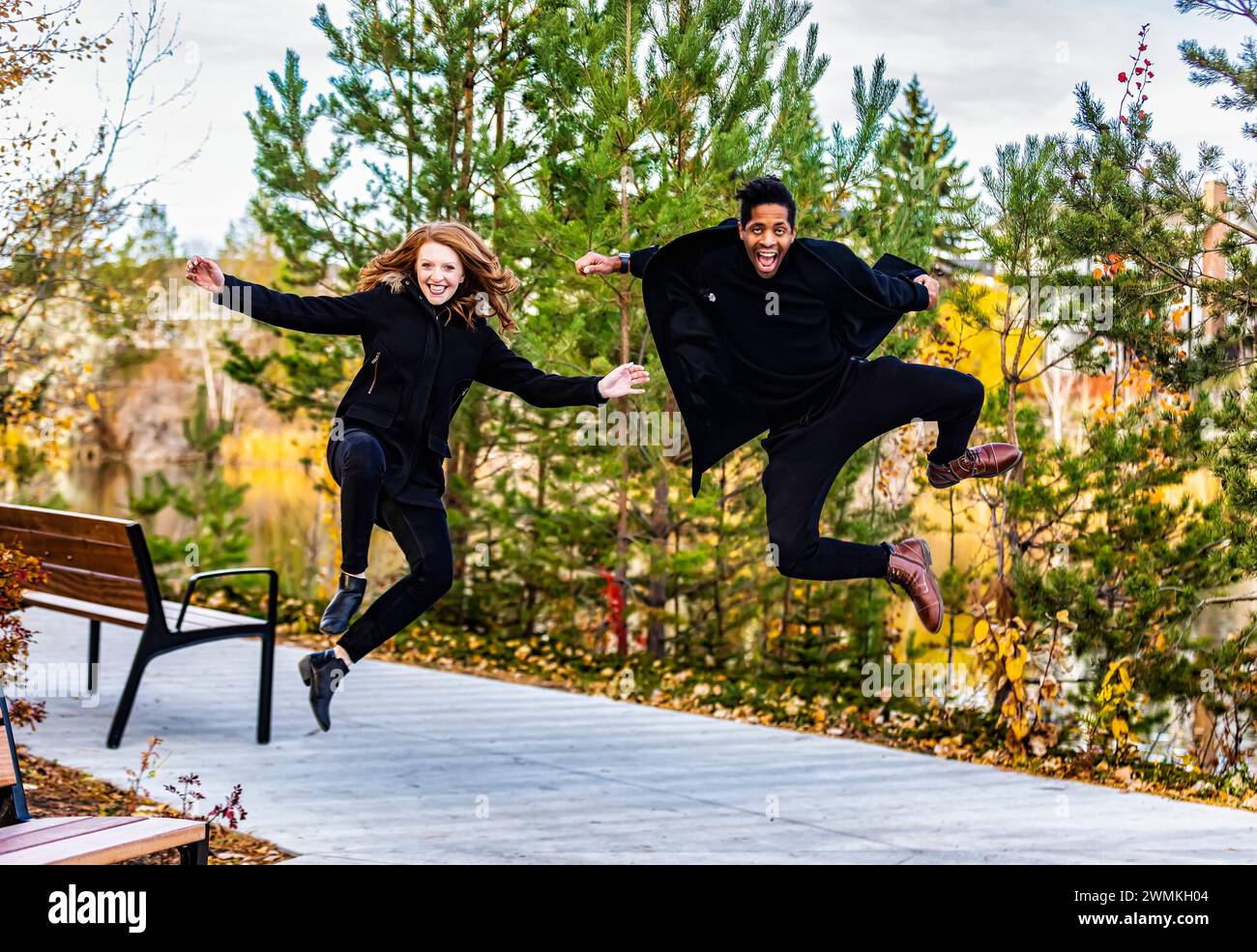 Portrait d'un couple de races mixtes sautant dans les airs, souriant à la caméra et passant du temps de qualité ensemble lors d'une sortie en famille d'automne dans un ci... Banque D'Images