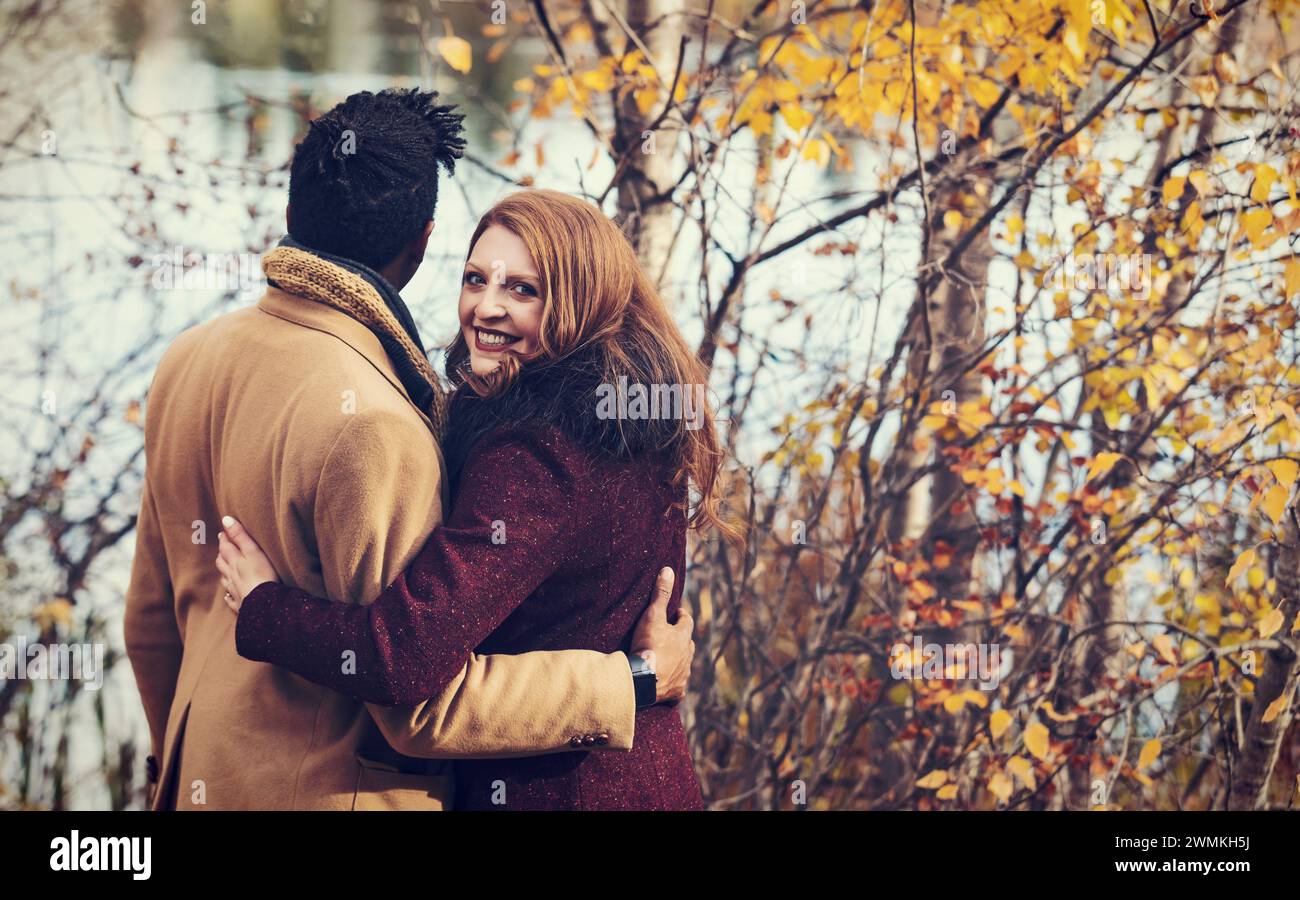 Vue prise de derrière un couple de races mixtes debout sur la rive d'un lac serrés l'un l'autre avec femme regardant en arrière et souriant à la caméra, ... Banque D'Images