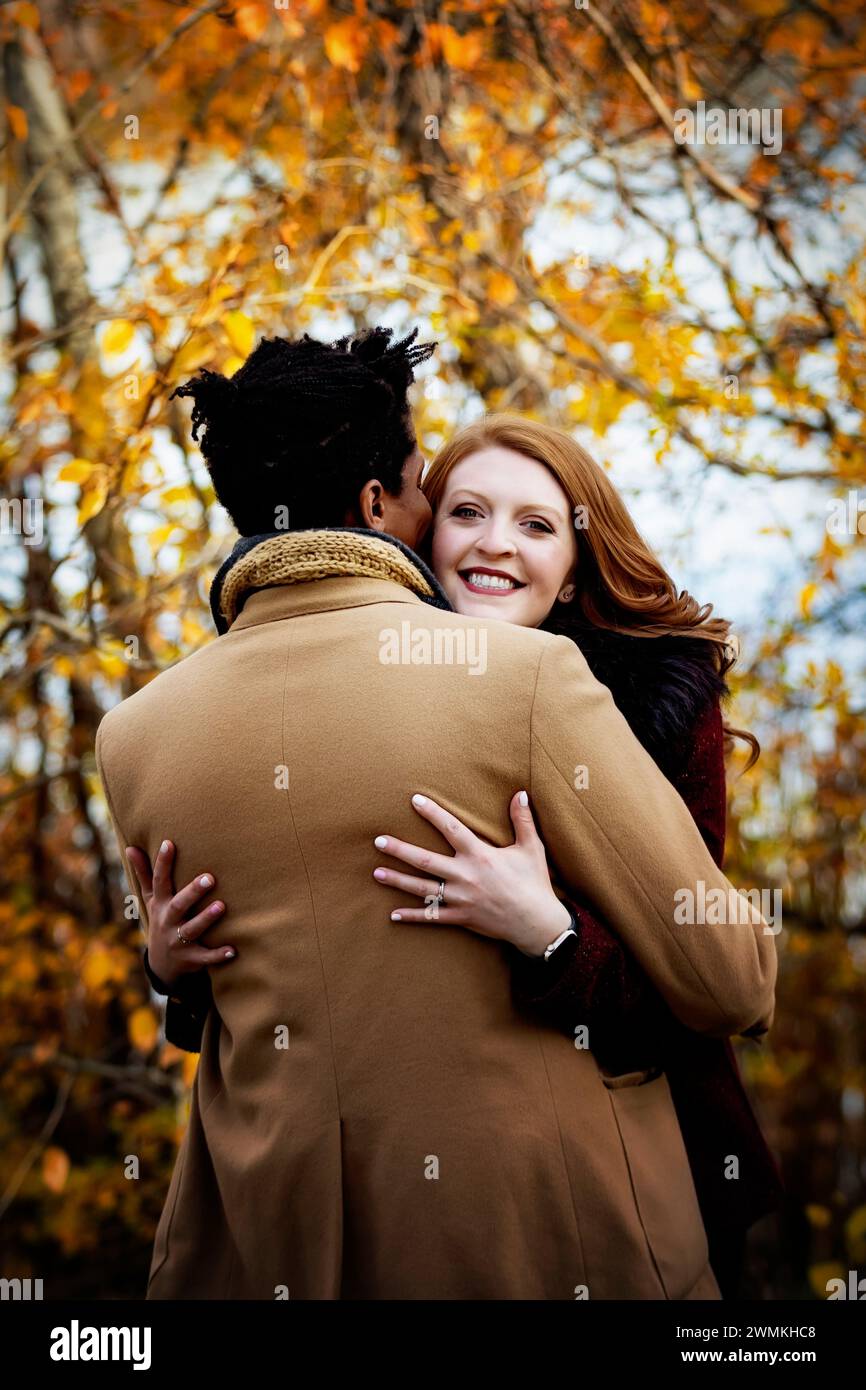 Vue prise de derrière d'un couple de races mixtes s'embrassant avec femme souriant à la caméra, passant du temps de qualité ensemble lors d'un automne fami... Banque D'Images