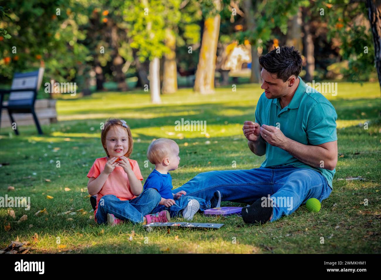 Père passant du temps de qualité et utilisant la langue des signes avec son jeune fils qui a le syndrome de Down, tandis que sa fille joue le long, dans un parc de la ville durin... Banque D'Images