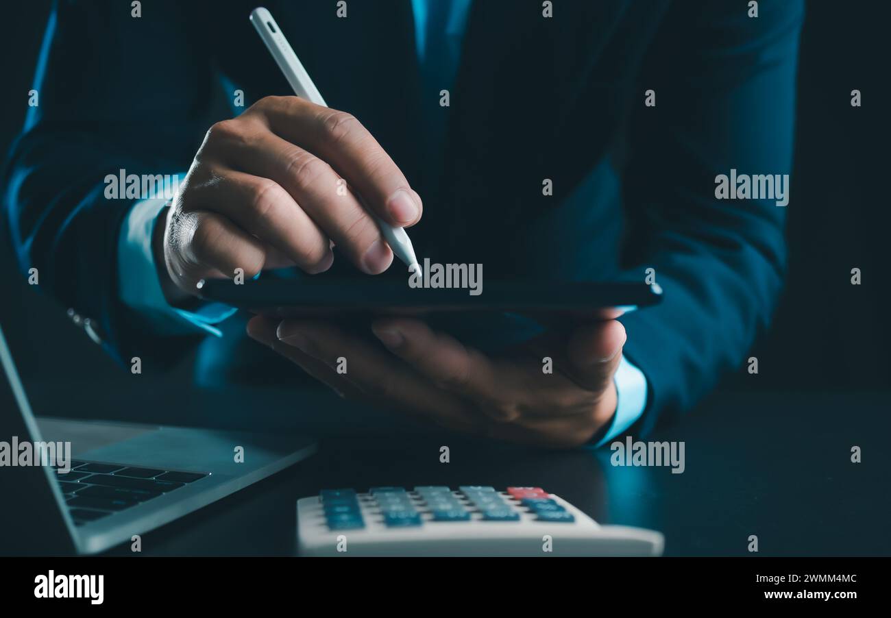 Homme d'affaires en costume noir travaillant sur ordinateur portable et tablette et calculatrice, mains toucher sur tablette au bureau avec fond sombre, travail en ligne Banque D'Images