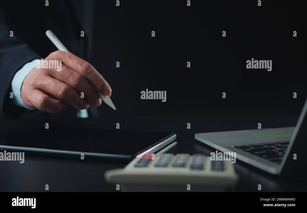 Homme d'affaires en costume noir travaillant sur ordinateur portable et tablette et calculatrice, mains toucher sur tablette au bureau avec fond sombre, travail en ligne Banque D'Images