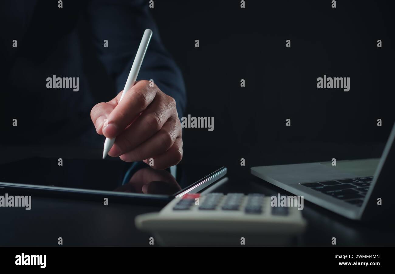 Homme d'affaires en costume noir travaillant sur ordinateur portable et tablette et calculatrice, mains toucher sur tablette au bureau avec fond sombre, travail en ligne Banque D'Images