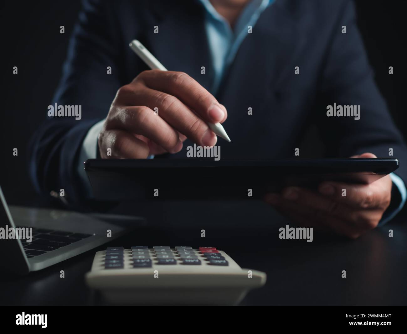 Homme d'affaires en costume noir travaillant sur ordinateur portable et tablette et calculatrice, mains toucher sur tablette au bureau avec fond sombre, travail en ligne Banque D'Images