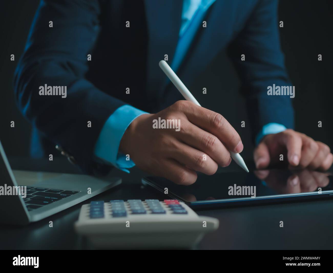 Homme d'affaires en costume noir travaillant sur ordinateur portable et tablette et calculatrice, mains toucher sur tablette au bureau avec fond sombre, travail en ligne Banque D'Images