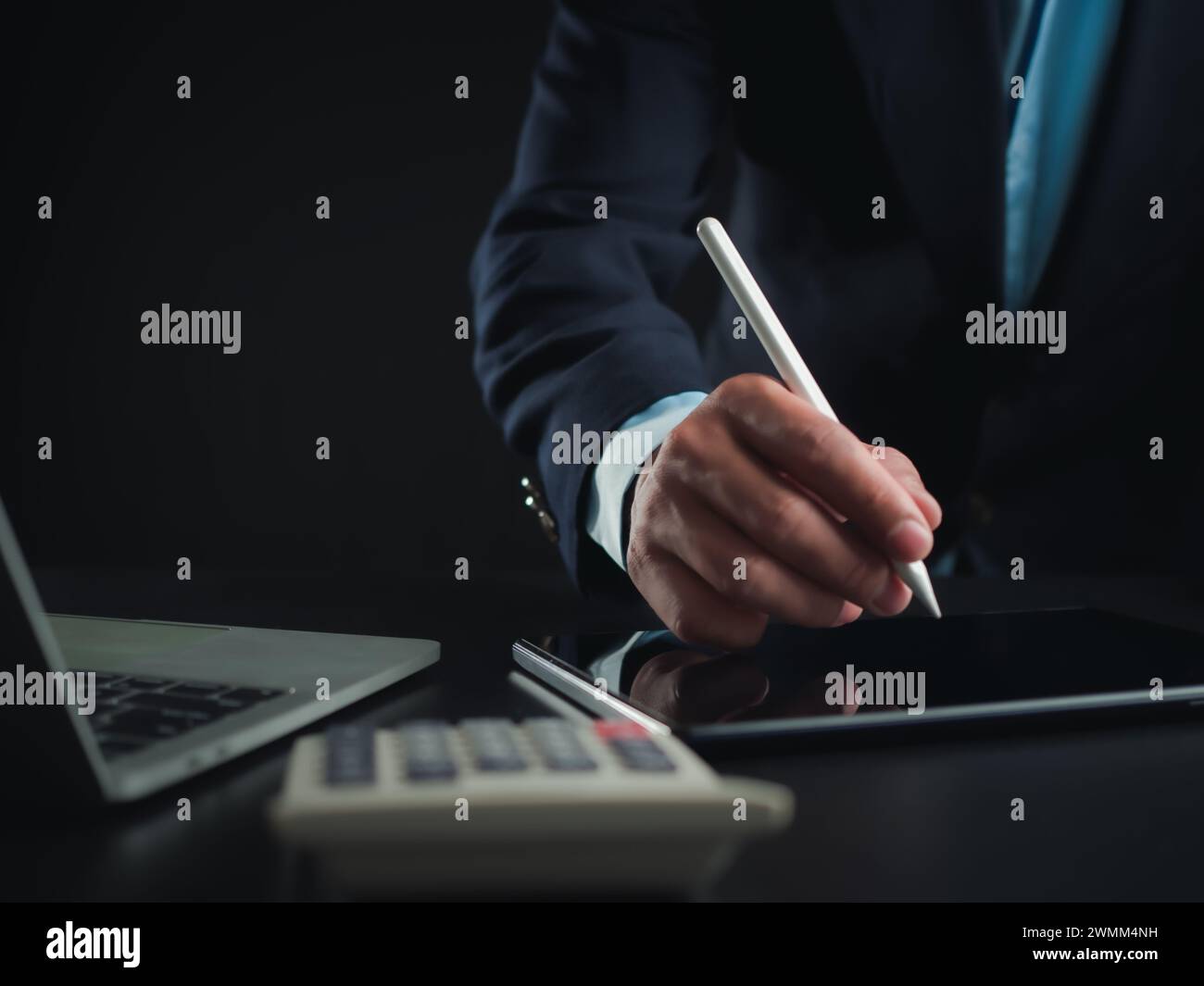 Homme d'affaires en costume noir travaillant sur ordinateur portable et tablette et calculatrice, mains toucher sur tablette au bureau avec fond sombre, travail en ligne Banque D'Images