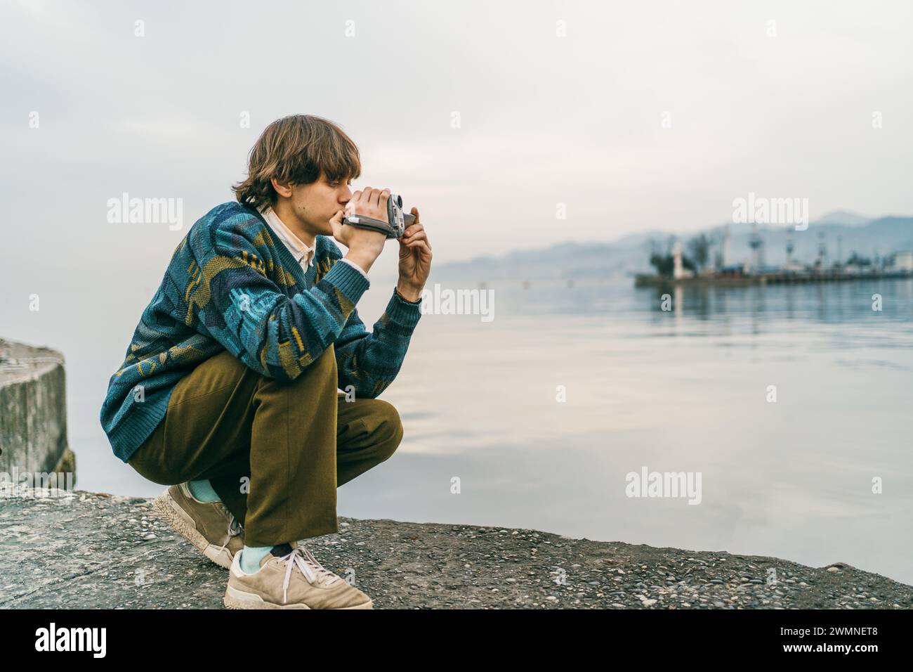 Réfléchi jeune homme accroupie par le lac tranquille ou la surface de la mer, capturant la scène avec la caméra rétro. Banque D'Images
