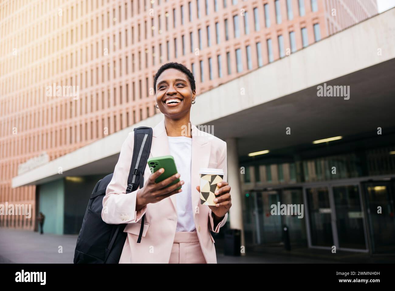 Jeune belle femme d'affaires avec un sac à dos qui va travailler le matin avec une tasse réutilisable et SMS sur un téléphone vert. Banque D'Images