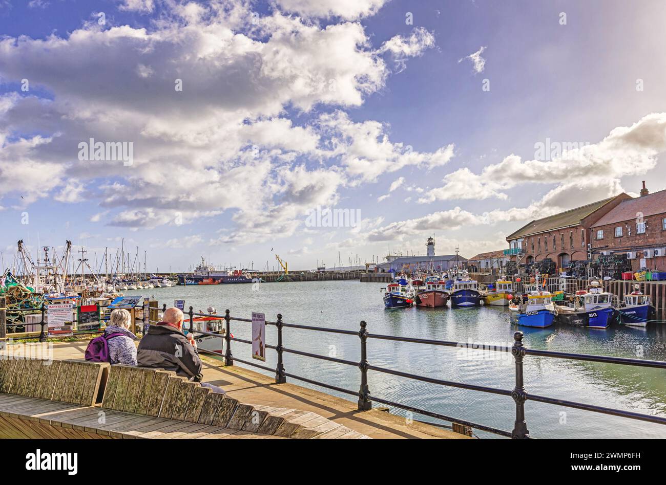 Les bateaux de pêche sont amarrés dans le port. Un quai est d'un côté et un phare est au loin. Un couple est assis à manger des glaces. Banque D'Images