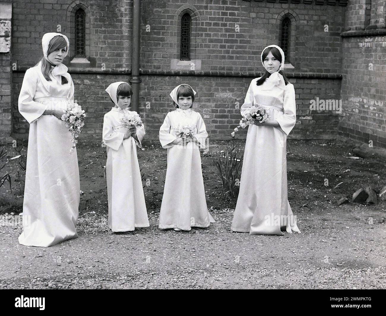 Années 1960, historique, quatre demoiselles d'honneur, deux jeunes femmes et deux petites filles debout dans leurs robes longues assorties et leurs chapeaux sur un chemin à l'extérieur d'une église, Angleterre, Royaume-Uni. Banque D'Images