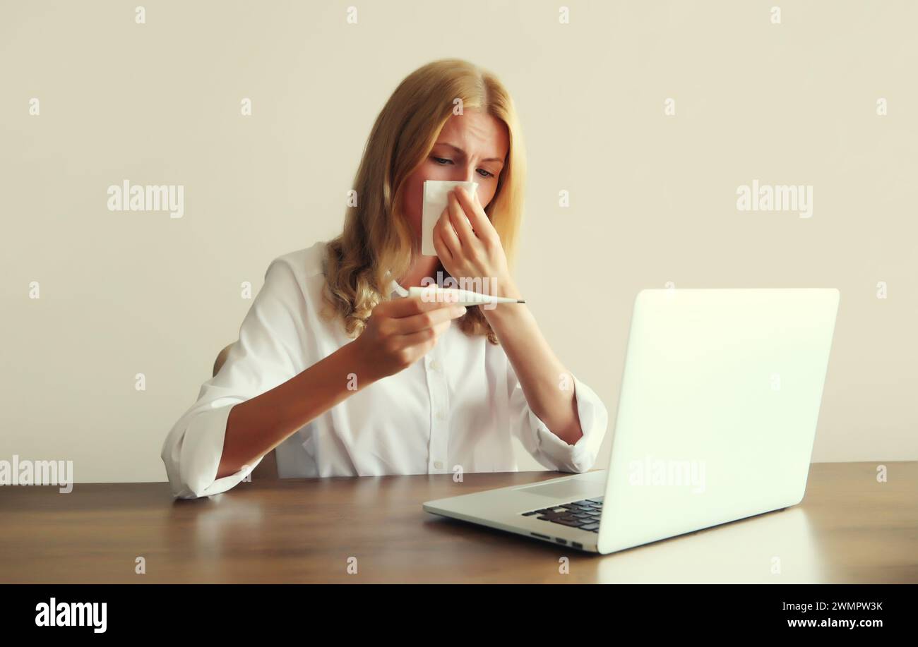 Femme malade éternuant nez de soufflage à l'aide de tissu, mesurant la température corporelle en vérifiant le thermomètre travaillant avec un ordinateur portable assis au bureau dans le bureau Banque D'Images