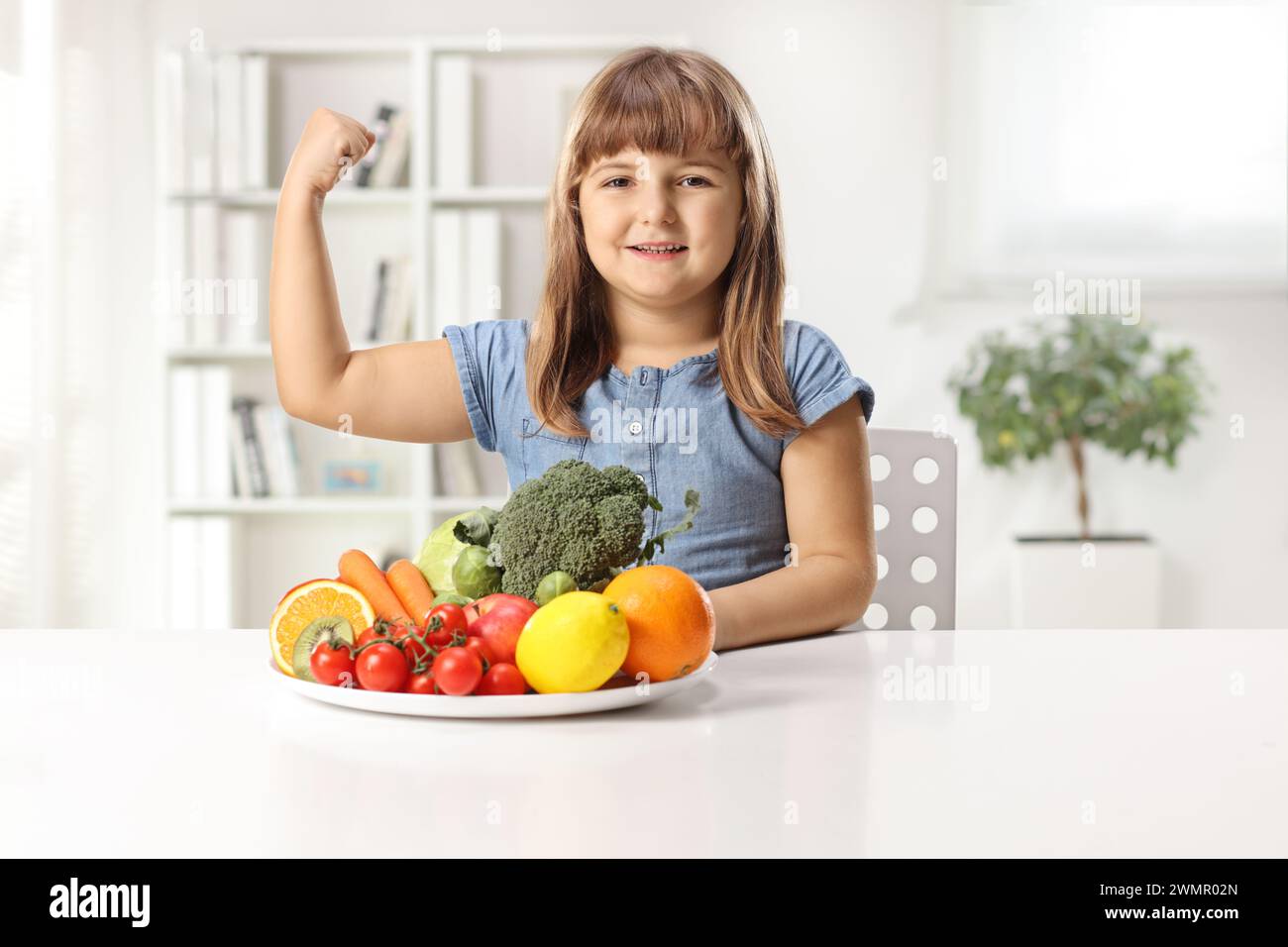 Forte petite fille assise avec une assiette de fruits et légumes sur une table et montrant du muscle Banque D'Images