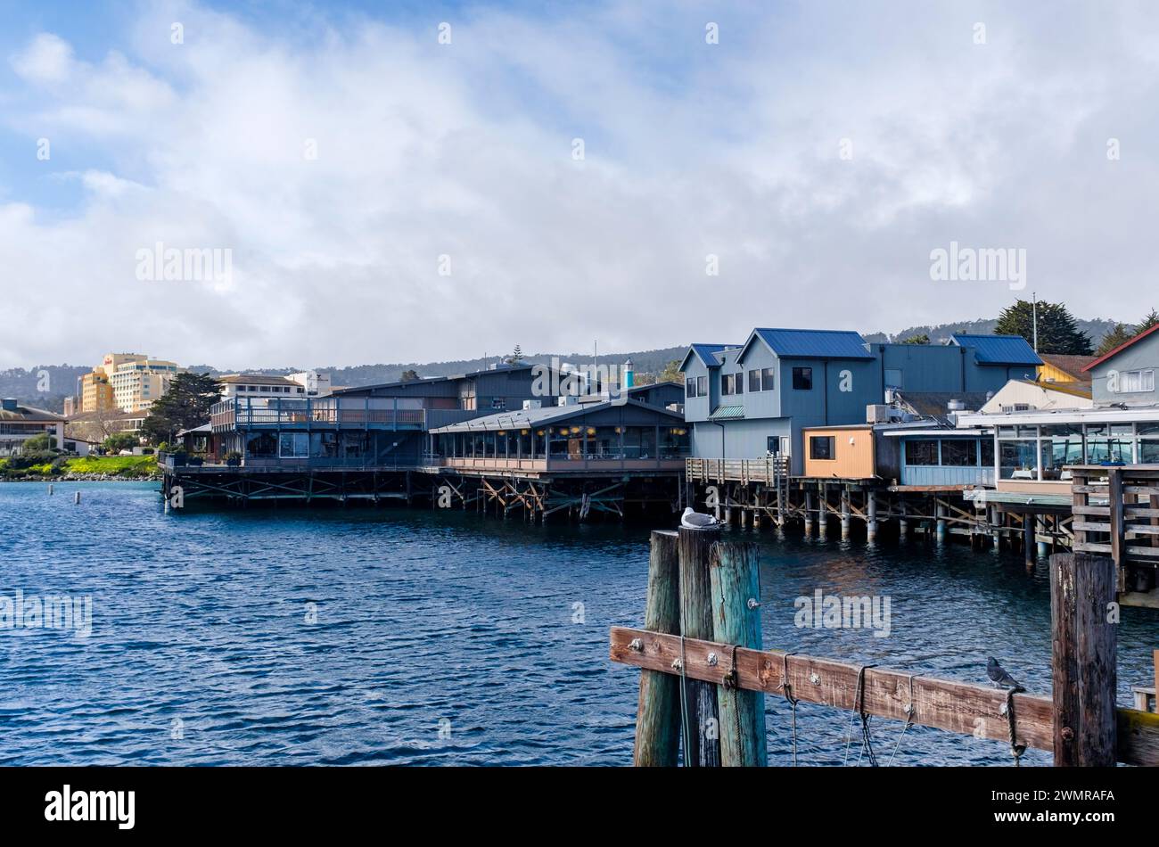 Des nuages d'ambiance atmosphériques survolent l'ancien quai des pêcheurs de Monterey où les restaurants et les boutiques surplombent le port. Une mouette repose sur une pile de bois. Banque D'Images