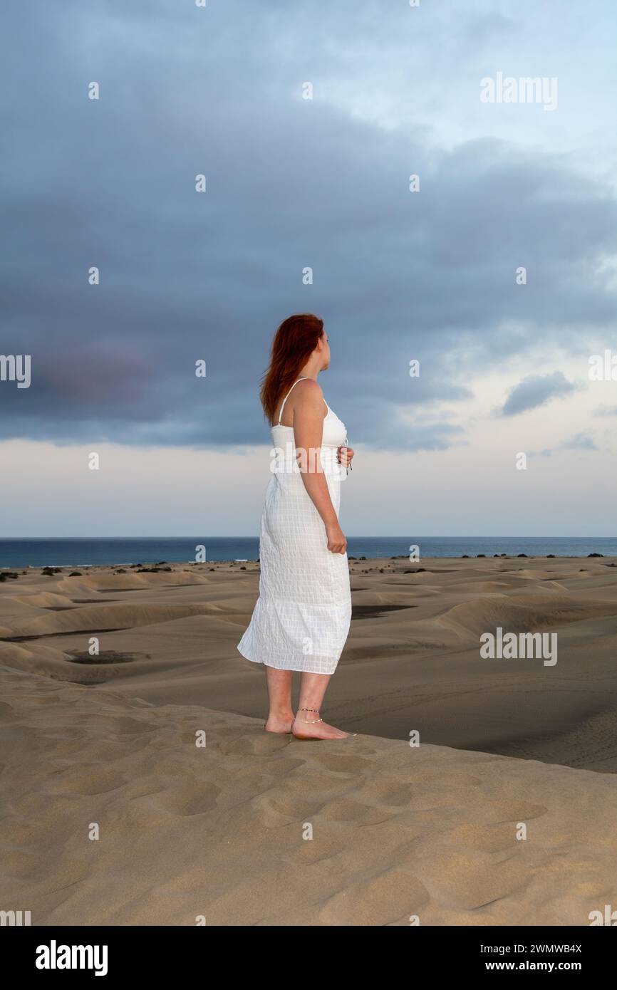 Une jeune fille se tient dans une robe blanche dans les dunes de sable avec le ciel et les nuages en fin d'après-midi Banque D'Images