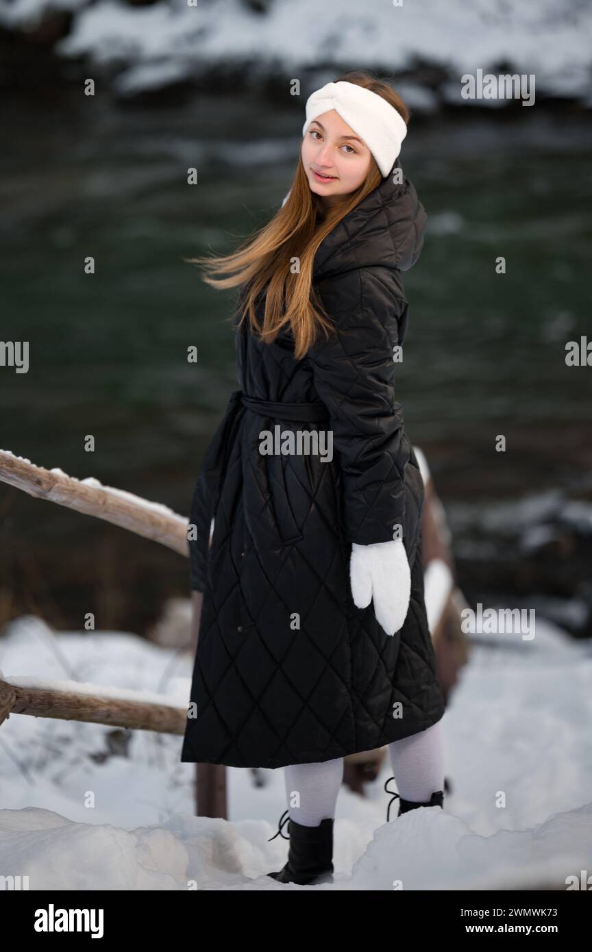 Belle fille en vêtements d'hiver dans la forêt près de la rivière en hiver, bandeau blanc d'hiver et gants blancs. Banque D'Images