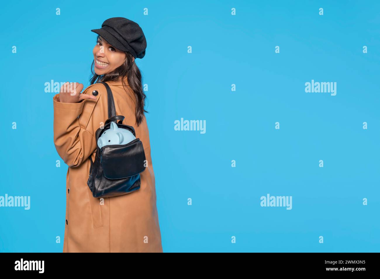 Heureuse femme indienne dans un manteau brun économiser de l'argent pour un jour de pluie dans la tirelire dans son sac à dos, investissement, économiser de l'argent, monnaie, dépôt. Intérieur Banque D'Images