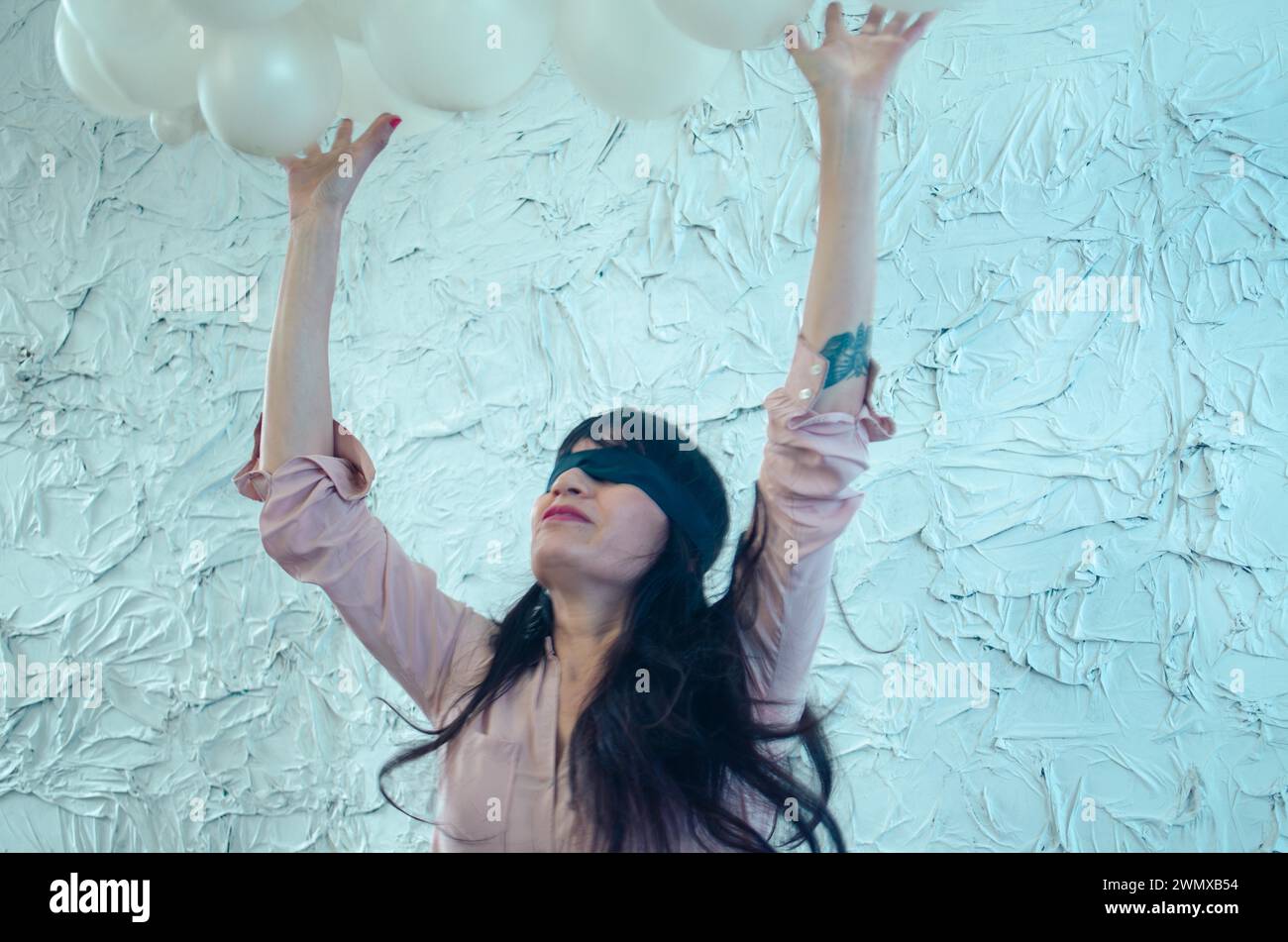 Une jeune femme hispanique adulte joueuse les yeux bandés atteignant vers le ciel, entourée d'une abondance de ballons blancs, performance artistique, danse ludique Banque D'Images