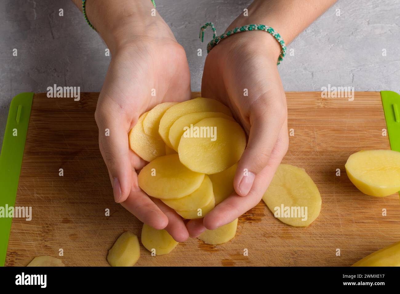 Les mains féminines authentiques tiennent des pommes de terre de coupe sur une planche à découper en bois sur la table de cuisine. Femme dans le tablier de pomme de terre coupé, préparation de la nourriture, intérieur de la maison, Banque D'Images