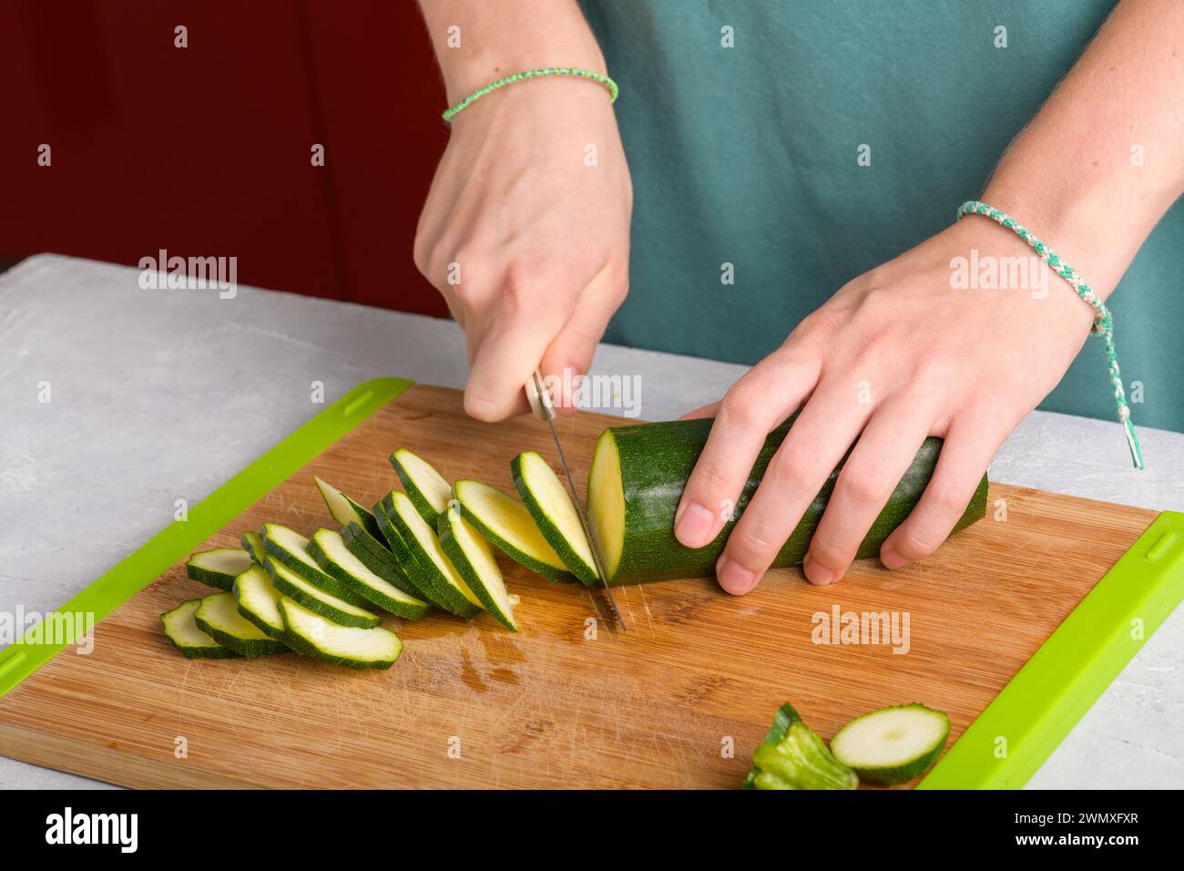 Mains féminines authentiques coupant les courgettes sur une planche à découper en bois sur la table de cuisine. Femme dans le tablier coupé courgettes pour préparer la nourriture. Végétalien et végétarien Banque D'Images
