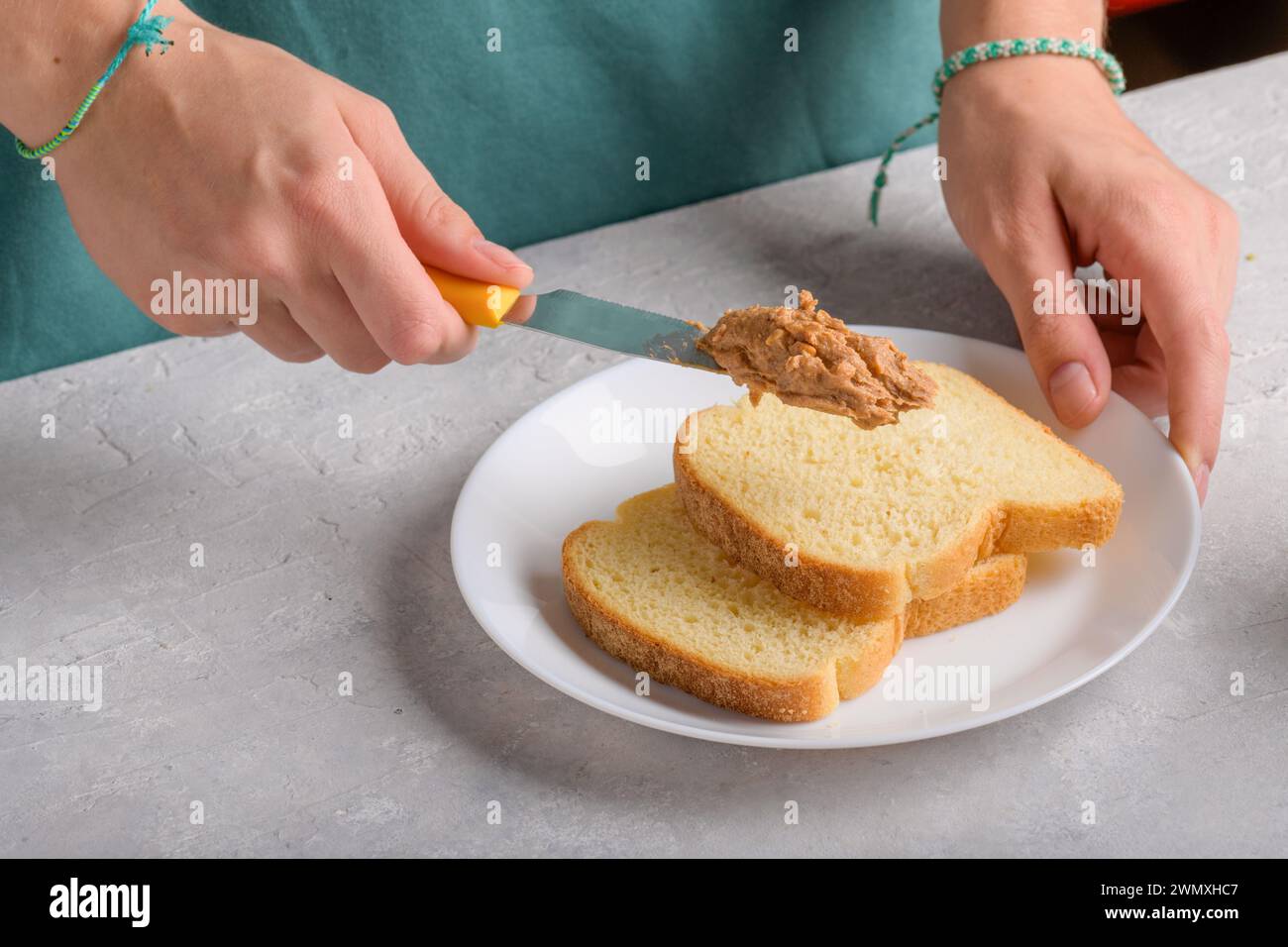 Mains féminines authentiques étalant du beurre d'arachide sur un morceau de pain blanc toast, à la table de cuisine grise, gros plan. Snack typique, style de vie alimentaire, dom Banque D'Images