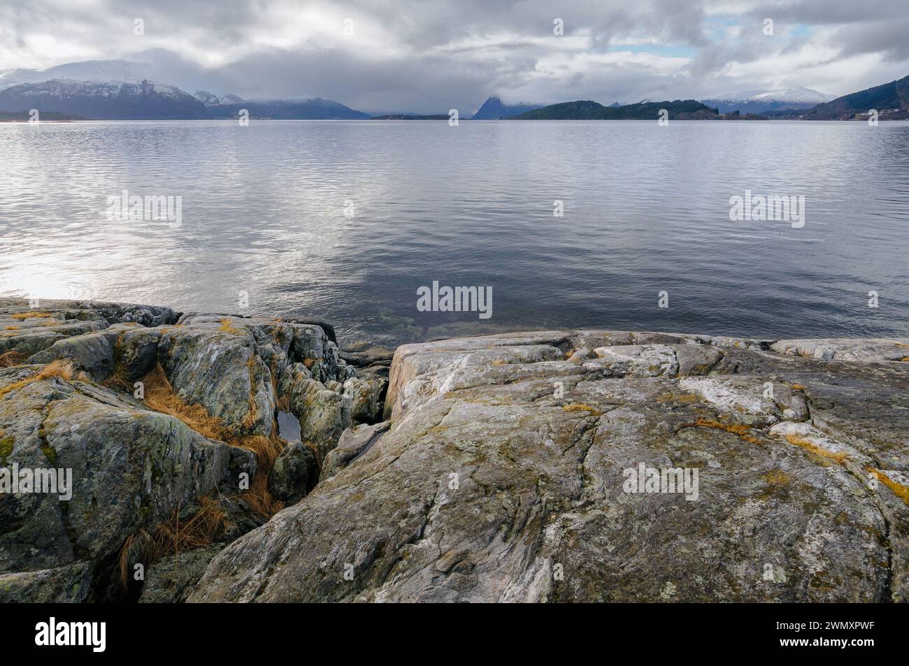 L'eau calme d'un fjord s'étend à l'horizon, bordée par une rive rocheuse et des montagnes lointaines sous un ciel nuageux. Banque D'Images