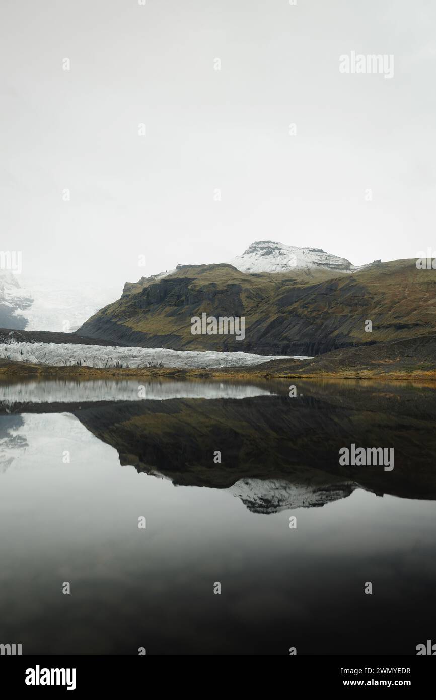 Une vue islandaise sereine avec le reflet d'un glacier dans des eaux tranquilles, entouré de montagnes brumeuses et de tons terreux. Banque D'Images