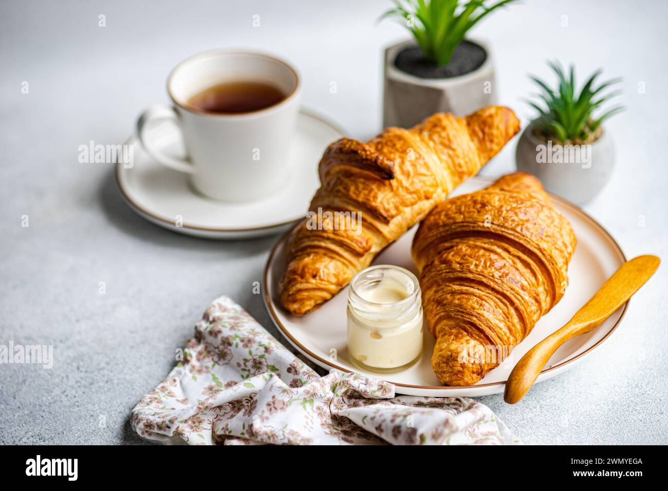 Un petit déjeuner avec deux croissants dorés sur une assiette, accompagné d'un pot de miel à la crème et une tasse de thé, avec de petites plantes en pot dans le dos Banque D'Images