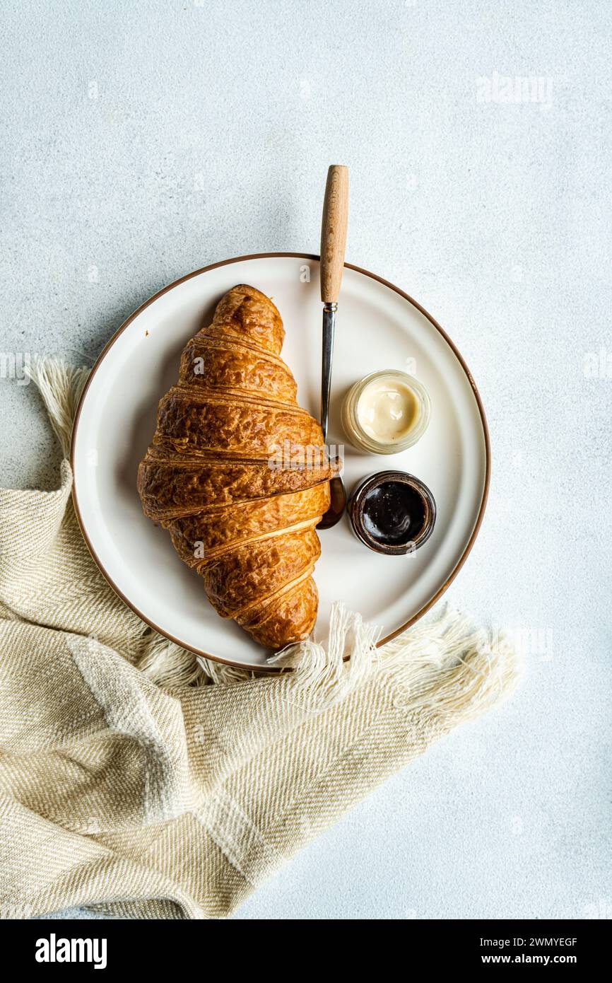 Vue de dessus de l'élégante assiette de petit déjeuner avec un croissant, des pots de miel de citron et de gingembre et de pâte à tartiner aux noisettes, accompagné d'un couteau sur un tissu tissé Banque D'Images