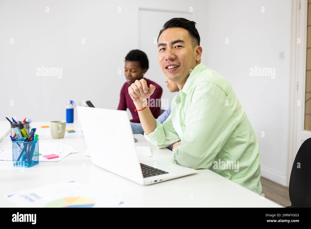 Un homme asiatique souriant dans une chemise verte décontractée participe à une réunion de bureau avec des collègues, créant une atmosphère de travail dynamique et collaborative. Banque D'Images