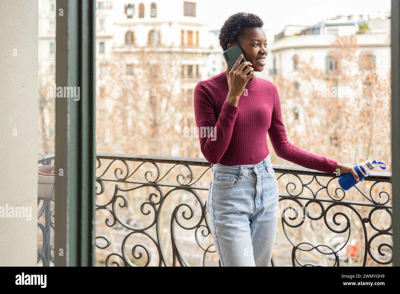 Jeune femme noire en tenue décontractée parlant sur un smartphone tout en se tenant debout sur un balcon avec toile de fond urbaine. Banque D'Images