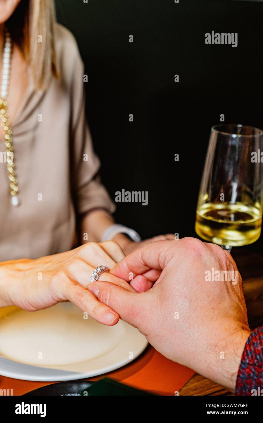 Gros plan de la main d'un homme plaçant une bague de fiançailles sur le doigt d'une femme lors d'un dîner romantique, avec un verre à vin en arrière-plan Banque D'Images