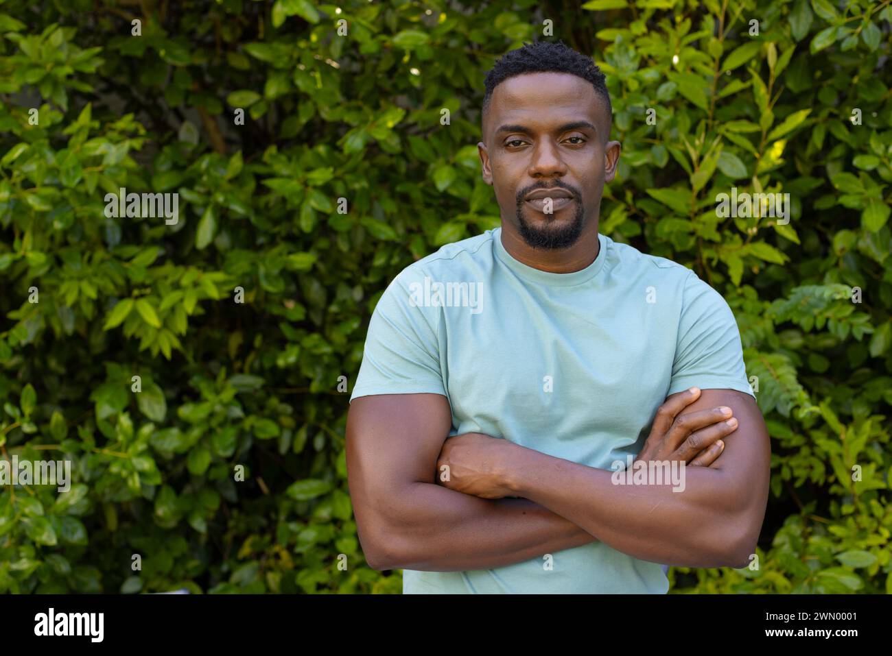 Homme afro-américain avec les bras croisés se tient confiant devant une haie verte luxuriante Banque D'Images