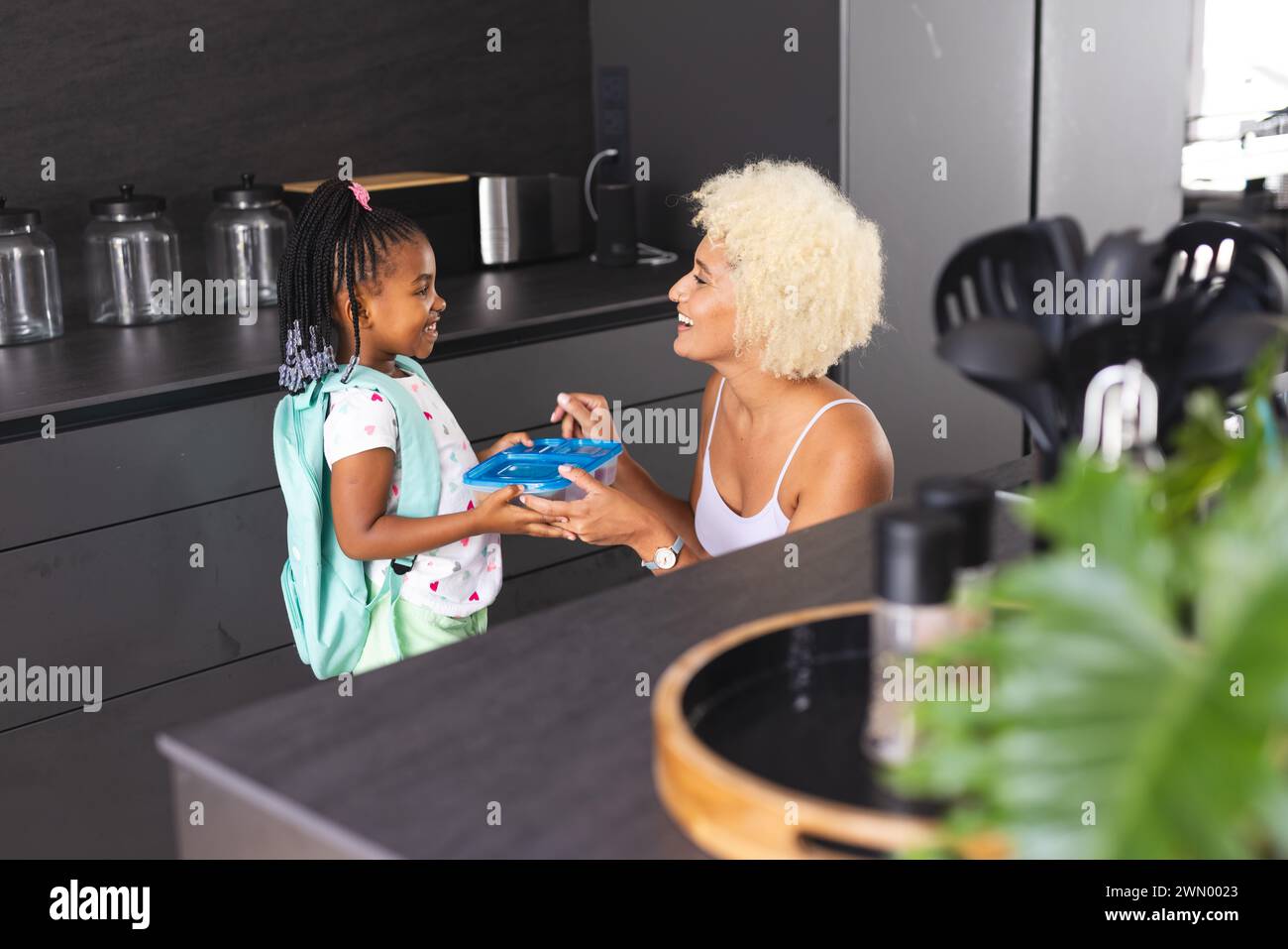 Une jeune mère biraciale aux cheveux blonds courts tend une boîte à lunch bleue à une fille afro-américaine Banque D'Images