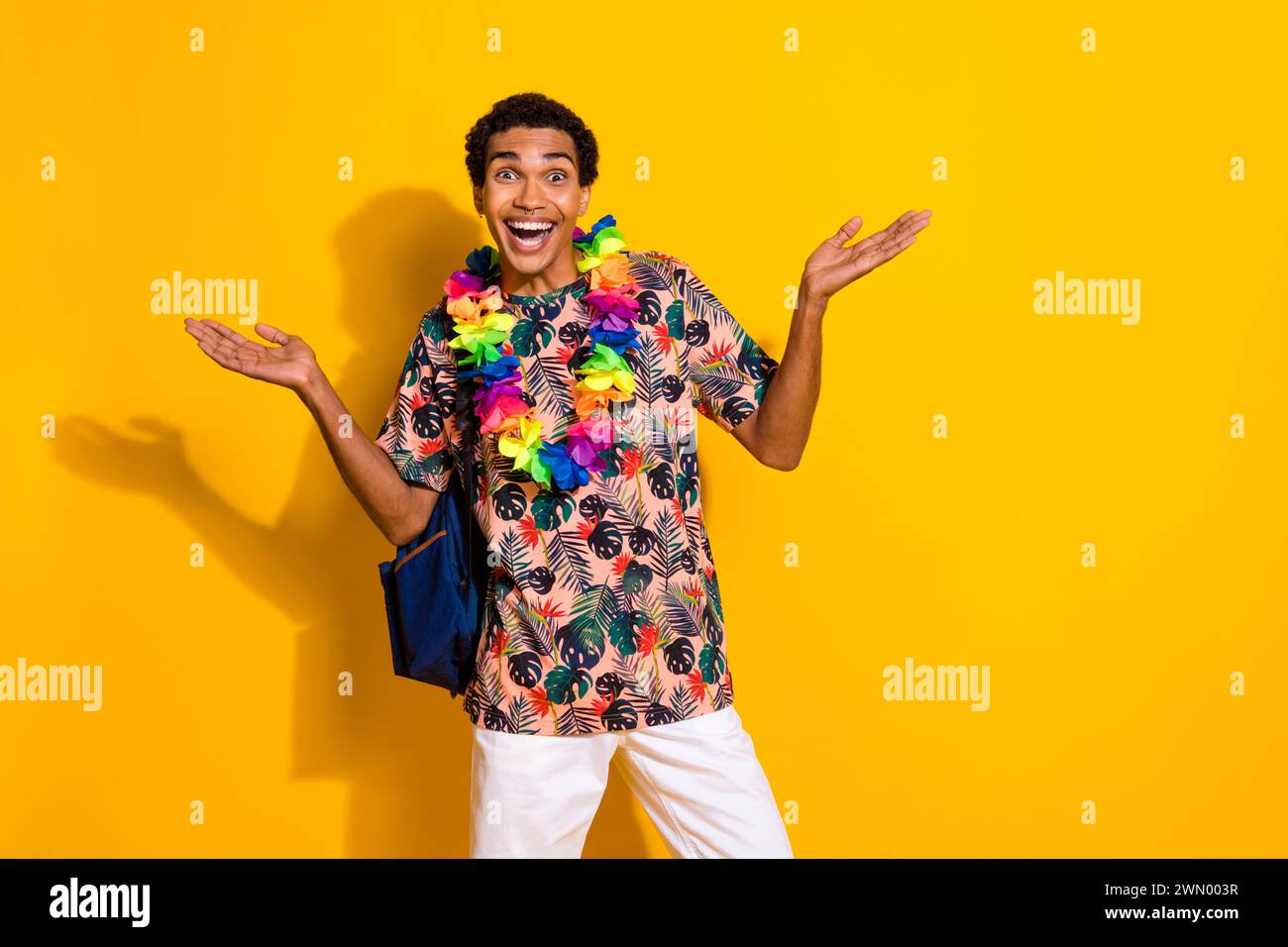 Photo de jeune gars positif funky dans le collier de fleurs de style exotique et sac à dos voyageur touristique isolé sur fond de couleur jaune Banque D'Images