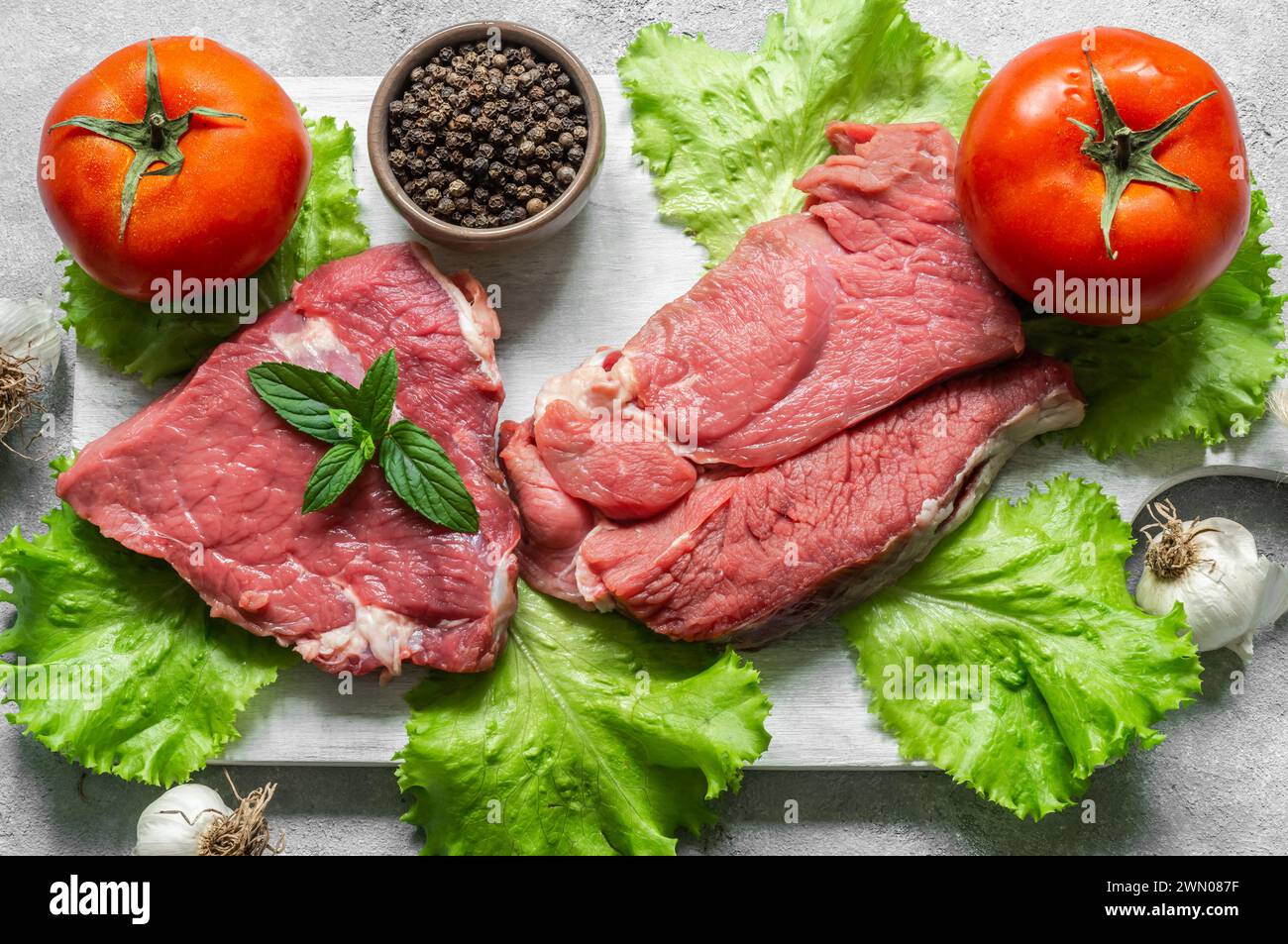 Viande de steak de bœuf crue sur planche à découper sur fond rustique avec des feuilles de menthe, des poivrons et de la laitue, concept de boucher Banque D'Images