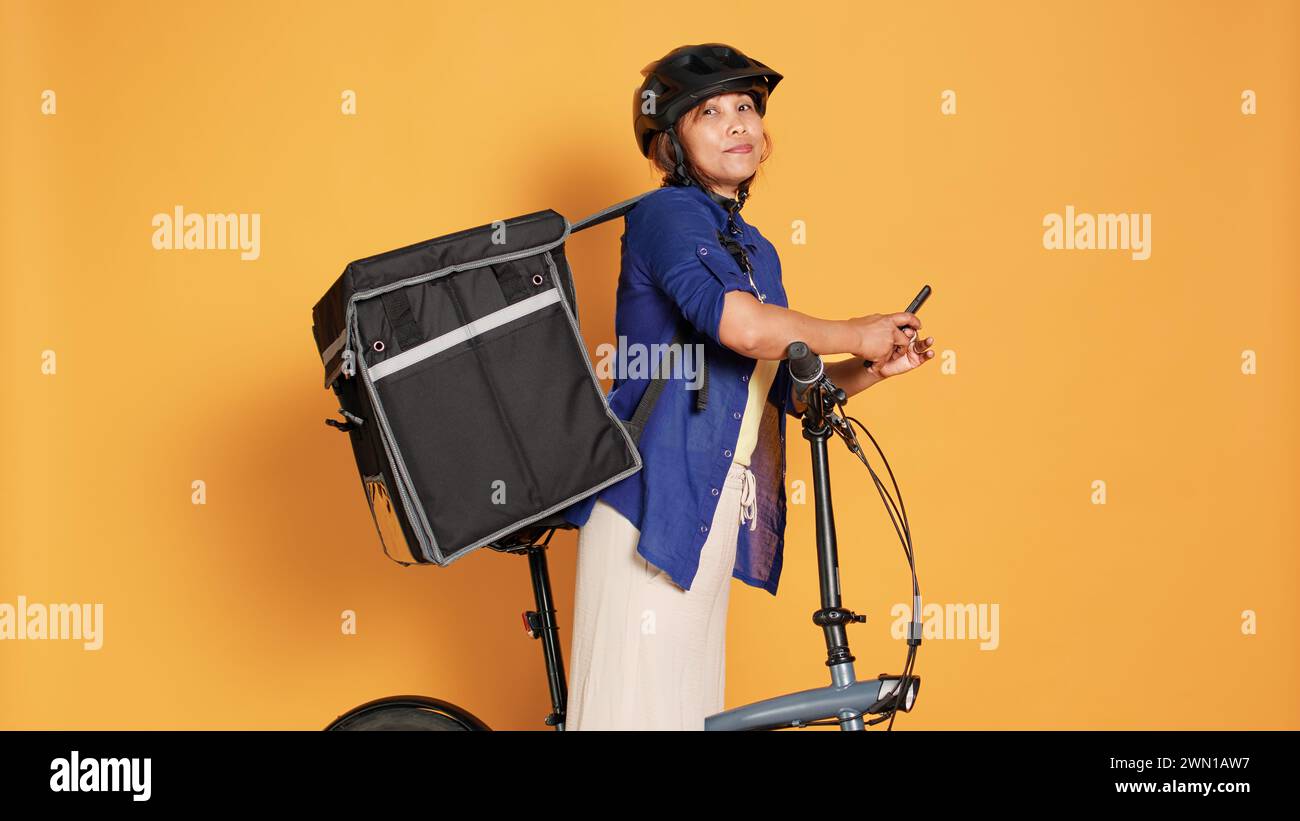 Femme de messagerie du BIPOC prenant une pause entre les livraisons de repas à emporter, envoyant des SMS à des amis. Cycliste attendant de recevoir la prochaine commande de nourriture du client, isolé sur fond orange de studio Banque D'Images