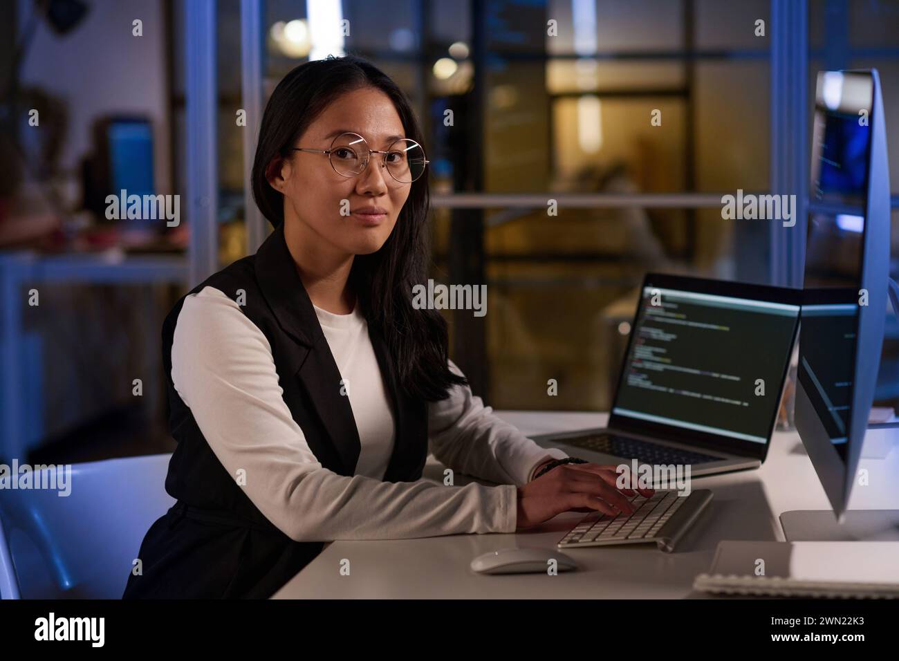 Portrait moyen d'une jeune programmeuse asiatique travaillant sur du code à l'aide d'un ordinateur portable et d'un ordinateur de bureau sur le lieu de travail dans le bureau de l'entreprise INFORMATIQUE tard dans la soirée Banque D'Images