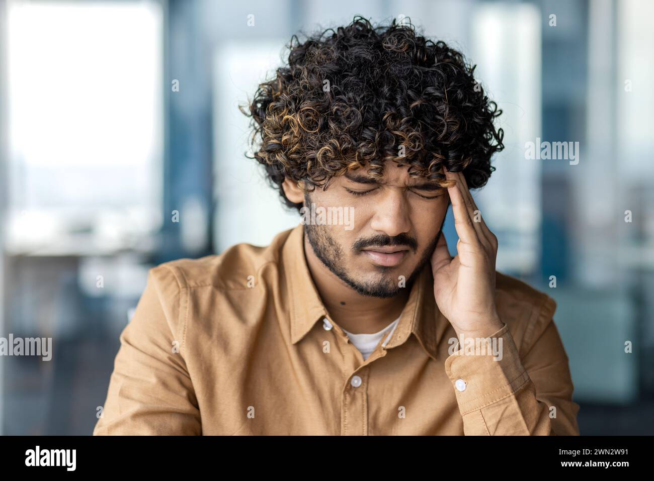 Portrait d'un musulman barbu en chemise fermant les yeux et massant le temple avec la main sur fond flou. Homme bouclé fatigué éprouvant des maux de tête au travail tout en se sentant dépassé par les délais. Banque D'Images