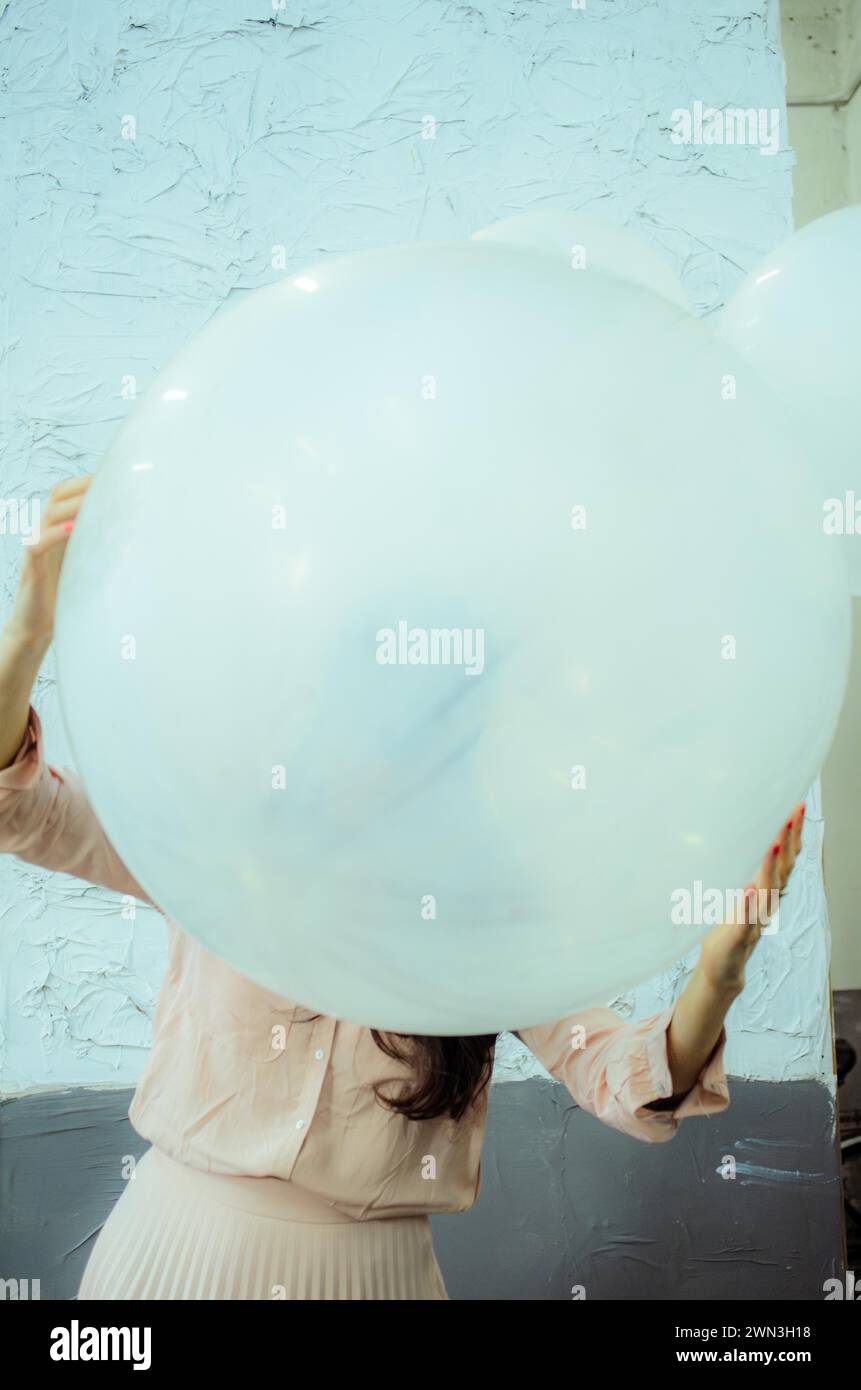 Femme ludique cachée derrière un grand ballon blanc, avec un fond de mur texturé, performance artistique, danse ludique Banque D'Images