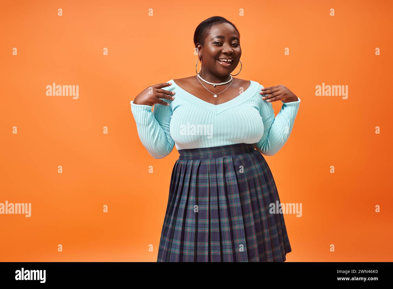 femme afro-américaine de plus grande taille heureuse en jupe à carreaux et manches longues bleues sur fond orange Banque D'Images