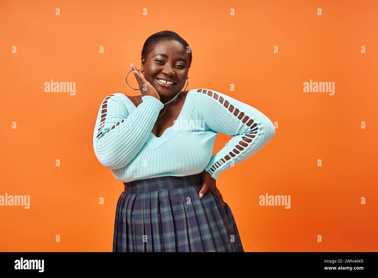 body positive femme afro-américaine en jupe à carreaux et manches longues bleues souriant sur orange Banque D'Images