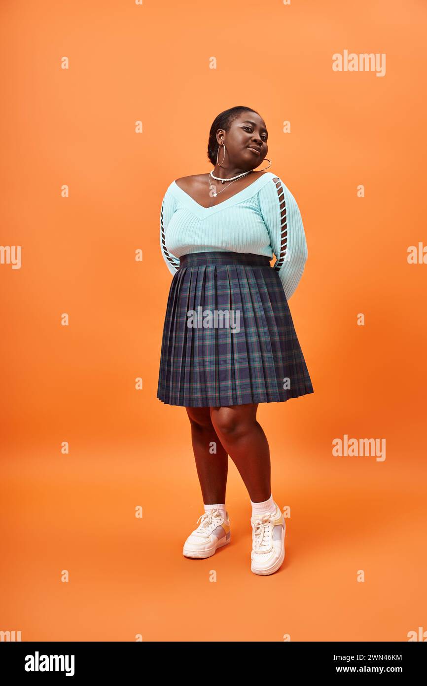 jeune femme afro-américaine de taille plus en jupe à carreaux et manches longues bleues sur fond orange Banque D'Images
