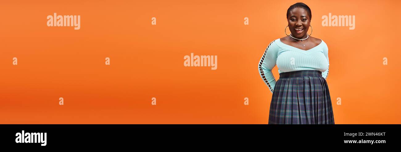 body positive femme afro-américaine en jupe à carreaux et manches longues bleues souriant sur orange, bannière Banque D'Images