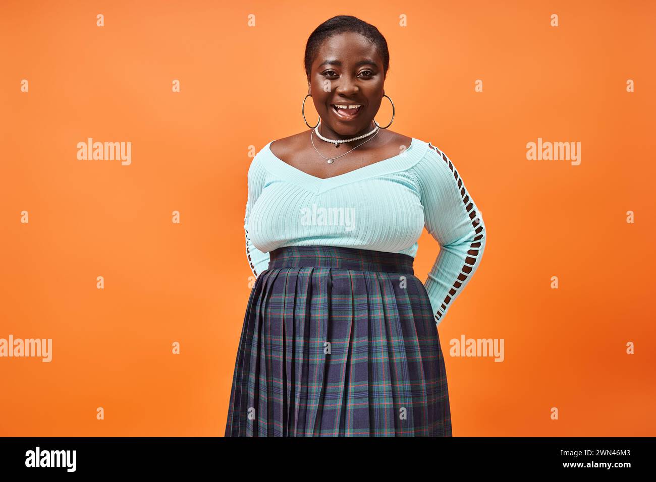 body positive femme afro-américaine en jupe à carreaux et manches longues bleues souriant sur fond orange Banque D'Images