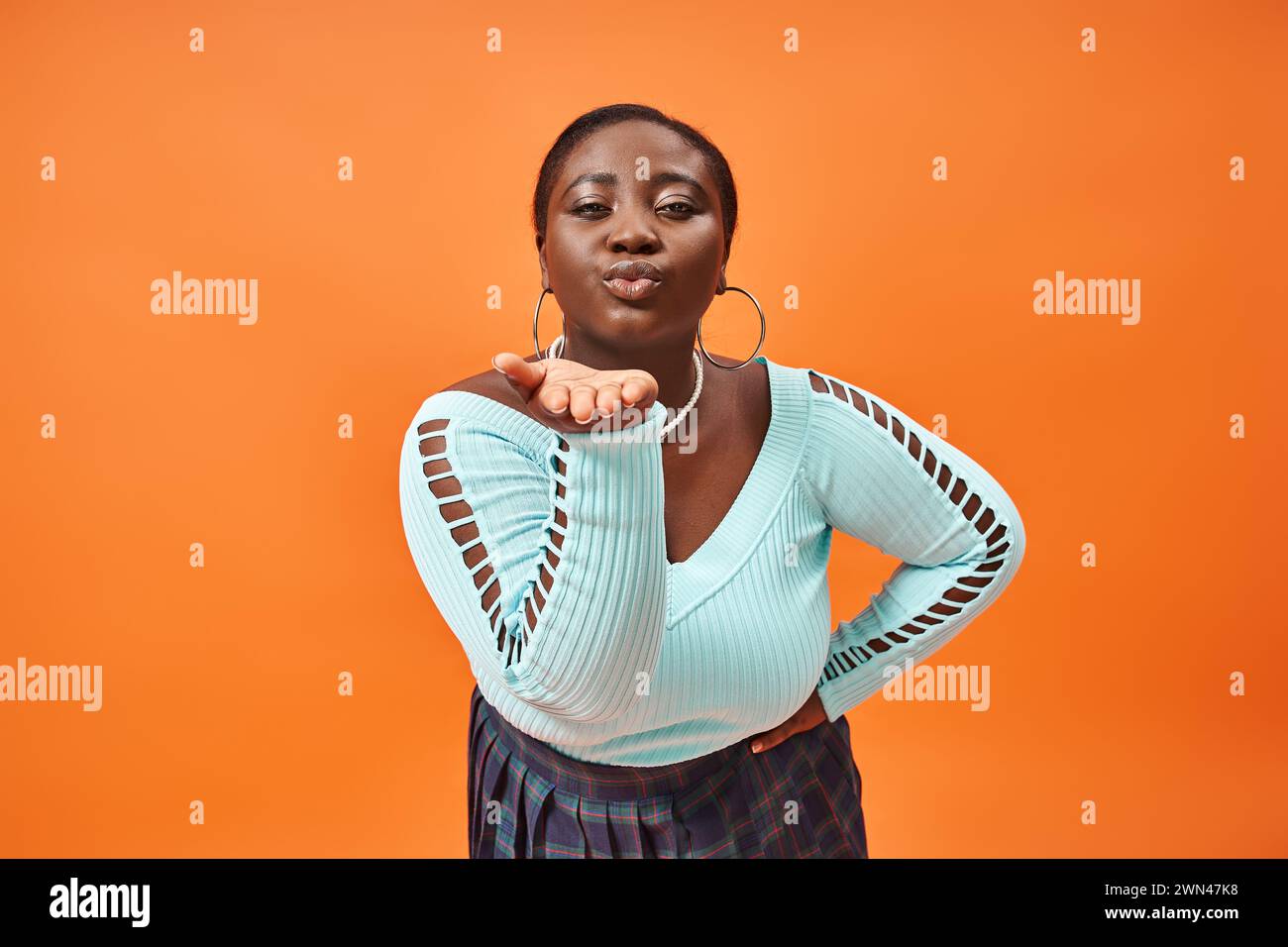 femme afro-américaine de taille plus en jupe à carreaux et manches longues envoyant un baiser aérien sur orange Banque D'Images